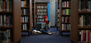 Vista frontal de una joven estudiante asiática usando un turbante usando una tableta y estudiando en una biblioteca - foto de stock