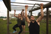 Front view of a young Caucasian man and a young Caucasian woman hanging from monkey bars at an outdoor gym during a bootcamp training session, with another participant also hanging from the bars in the background — Stock Photo