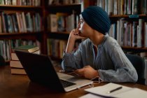 Бічний вид на юну азіатську студентку, яка носить тюрбан за допомогою ноутбука і навчається в бібліотеці — стокове фото