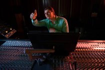 Передній погляд молодого кавказького звукоінженера, який сидів і працював за столом змішування в студії звукозапису, під час запису даючи пальцями вгору знак. — стокове фото