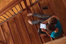 Vista aérea de dos niños caucásicos preadolescentes sentados en una escalera en casa, usando una tableta y un teléfono inteligente - foto de stock