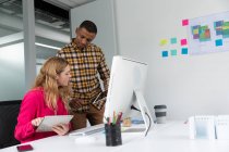 Vue latérale d'un jeune homme afro-américain debout et d'une jeune femme caucasienne assise à un bureau à l'aide d'ordinateurs tablettes dans le bureau moderne d'une entreprise créative — Photo de stock