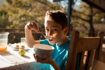 Porträt eines vorjugendlichen kaukasischen Jungen, der an einem Tisch sitzt und in einem Garten frühstückt und aus einer Schüssel isst — Stockfoto