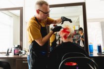 Бічний вид середнього віку Кавказький самець перукаря і молода кавказька жінка з яскраво-червоним волоссям в салоні волосся, з середнім віком змішана раса жіноча перукарня відображена в дзеркалі. — стокове фото