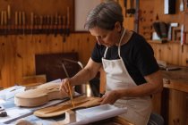 Seitenansicht einer älteren kaukasischen Geigenbauerin, die auf einer Werkbank in ihrer Werkstatt am Korpus einer Geige arbeitet, im Hintergrund hängen Werkzeuge an der Wand — Stockfoto