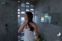 Vista laterale di una giovane donna bruna caucasica che indossa un asciugamano da bagno spazzolandosi i capelli in un bagno moderno — Foto stock