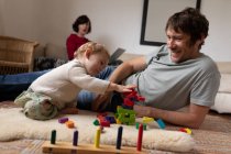 Vista frontal de perto de um jovem pai caucasiano brincando com seu bebê em um chão, enquanto uma jovem mãe caucasiana sentada no fundo — Fotografia de Stock