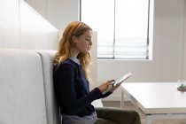 Seitenansicht einer jungen kaukasischen Frau, die mit einem Tablet-Computer im Essbereich eines kreativen Unternehmens auf einer Bank sitzt — Stockfoto