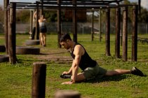 Vue latérale d'un jeune homme caucasien assis sur l'herbe s'étirant à une salle de gym extérieure avant une séance d'entraînement bootcamp — Photo de stock