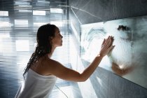 Вид збоку молодої кавказька жінка-брюнетка витираючи пар від дзеркала, стоячи у сучасній ванній — стокове фото