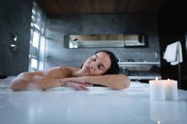 Retrato de una joven morena caucásica sentada en un baño de espuma con velas encendidas en el borde, apoyada en un costado y descansando con los ojos cerrados - foto de stock