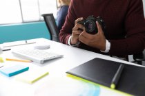 Вид спереду посередині чоловіка, що сидить за столом, тримає камеру SLR в сучасному офісі творчого бізнесу — стокове фото