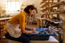 Vista lateral de uma jovem cerâmica caucasiana sentada e trabalhando com argila em uma roda de oleiros em um estúdio de cerâmica — Fotografia de Stock