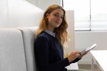 Бічний вид на молоду кавказьку жінку, яка сидить на лавці сидячи за допомогою планшетного комп'ютера в їдальні творчого бізнесу, звертаючись і посміхаючись до камери. — стокове фото