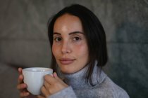 Ritratto da vicino di una giovane donna bruna caucasica che guarda la telecamera sorridente e tiene in mano una tazza di caffè — Foto stock
