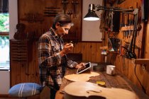 Вид збоку старшого Кавказького жіночого luthier за допомогою планшетного комп'ютера з інструментами звисають на стіні у фоновому режимі — стокове фото