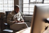 Nahaufnahme eines jungen afrikanisch-amerikanischen Mannes, der an einem Schreibtisch am Fenster sitzt und sein Smartphone in einem kreativen Büro benutzt — Stockfoto