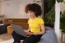 Вид спереди на молодую женщину смешанной расы, сидящую на бобовом мешке со скрещенными ногами с помощью ноутбука в креативном офисе — стоковое фото
