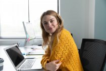 Porträt einer jungen kaukasischen Frau, die sich mit einem Laptop an einem Schreibtisch im modernen Büro eines kreativen Unternehmens umdreht und in die Kamera lächelt — Stockfoto