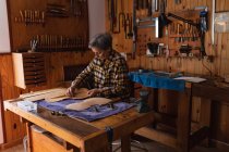 Seitenansicht einer älteren kaukasischen Geigenbauerin, die in ihrer Werkstatt am Korpus einer Geige arbeitet, im Hintergrund hängen Werkzeuge an der Wand — Stockfoto