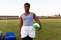 Портрет молодої дорослої змішаної раси жінки-регбіста, що стоїть на спортивному полі, що тримає м'яч регбі під час тренувальної сесії — стокове фото