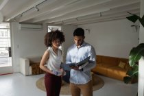 Вид спереду молодого змішаного чоловіка і молодої змішаної раси жінки, що стоїть, перевіряє документи і обговорює в творчому офісі — стокове фото