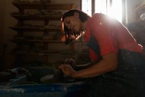 Seitenansicht einer lächelnden jungen kaukasischen Töpferin, die in einem Töpferatelier sitzt und mit Ton auf einer Töpferscheibe arbeitet, die von Sonnenlicht beleuchtet wird — Stockfoto