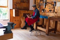 Seitenansicht einer älteren kaukasischen Geigenbauerin in einer Werkstatt mit einem Tablet-Computer, dessen Werkzeug im Hintergrund an der Wand hängt — Stockfoto