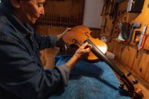 Seitenansicht einer älteren kaukasischen Geigenbauerin, die in ihrer Werkstatt eine Geige herstellt, im Hintergrund hängen Werkzeuge an der Wand — Stockfoto