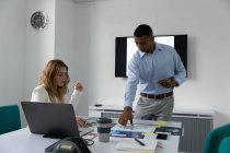 Front view of a young African American man standing holding a tablet computer and talking with a young Caucasian woman sitting at a desk, they are using a laptop computer and looking at visuals together in the modern office of a creative business — Stock Photo