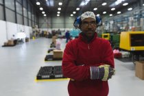 Ritratto ravvicinato di un operaio afro-americano di mezza età vestito in abiti da lavoro con paraorecchie in testa, in piedi in un magazzino di uno stabilimento di lavorazione che guarda alla telecamera con le braccia incrociate — Foto stock