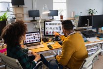 Nahaufnahme eines jungen Mannes mit gemischter Rasse und einer jungen Frau mit gemischter Rasse, die an einem Schreibtisch sitzen, auf einen Computerbildschirm schauen und in einem Kreativbüro diskutieren — Stockfoto