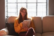 Vista frontale da vicino di una giovane donna caucasica seduta su un divano utilizzando un tablet nell'area salotto di un ufficio creativo, retroilluminata dalla luce del sole — Foto stock