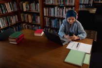 Vue de face d'une jeune étudiante asiatique portant un turban et un casque, utilisant un smartphone, un ordinateur portable et étudiant dans une bibliothèque — Photo de stock