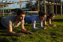 Vue latérale de deux jeunes femmes caucasiennes et d'un jeune homme caucasien faisant l'exercice de planche dans un gymnase extérieur lors d'une séance d'entraînement de bootcamp — Photo de stock