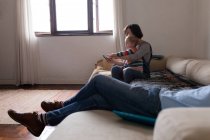 На вигляд, як молода кавказька мати сидить на дивані і користується смартфоном зі своєю дитиною, молодий батько лежить на дивані на передньому плані. — стокове фото