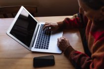Вид збоку крупним планом старший кавказька жінка сидить за столом вдома за допомогою портативного комп'ютера з смартфоном поруч з нею — стокове фото