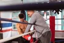 Side view close up of a young Caucasian male boxer by a boxing ring having his boxing gloves checked by a middle aged Caucasian male trainer — Stock Photo