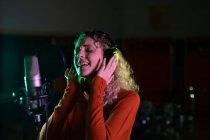 Side view close up of a young Caucasian female singer wearing headphones and singing in front of a microphone in a recording studio, with her eyes closed — Stock Photo