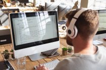 Visão traseira de perto de um jovem caucasiano sentado em uma mesa usando fones de ouvido e trabalhando em um computador em um escritório criativo — Fotografia de Stock