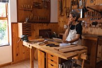 Side view of a senior Caucasian female luthier using a tablet computer and thinking in her workshop, with tools hanging up on the wall in the background — Stock Photo