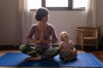 Front view of a young Caucasian mother sitting on a floor in a yoga pose next to her baby — Stock Photo