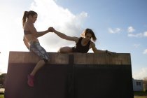 Seitenansicht von zwei jungen kaukasischen Frauen, die während eines Bootcamp-Trainings in einem Outdoor-Fitnessstudio über eine Wand klettern, eine sitzt oben und hilft der anderen nach oben — Stockfoto