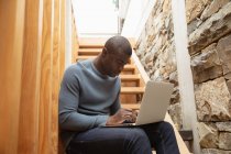 Вид сбоку на молодого афроамериканца с ноутбука, сидящего дома на лестнице — стоковое фото