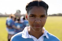 Portrait close up of a young adult mixed race female rugby player standing on a rugby pitch looking to camera, with her teammates in a huddle together in the background — Stock Photo