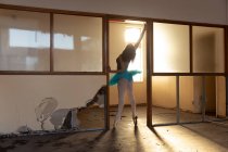 Вид спереду молодої змішаної раси жіночої балетної танцівниці в блакитному взутті, що танцює у дверному отворі в покинутій будівлі складів, підсвічування сонячним світлом — стокове фото