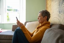 Вид сбоку на взрослую белую женщину с короткими седыми волосами в свитере, сидящую дома с помощью смартфона и улыбающуюся, с залитым солнцем окном на заднем плане — стоковое фото