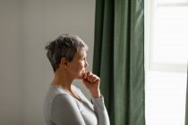 Vue latérale d'une femme blanche mature avec des cheveux gris courts debout et regardant par la fenêtre à la maison — Photo de stock