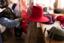 Feche a mão de uma mulher segurando um chapéu vermelho enquanto ela opera uma máquina para limpá-lo a vapor na oficina em uma fábrica de chapéus, outros chapéus visíveis no fundo — Fotografia de Stock