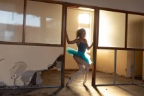 Frontansicht einer jungen Balletttänzerin mit gemischter Rasse, die ein blaues Tutu und Spitzenschuhe trägt und in einem Eingang eines verlassenen Lagergebäudes tanzt, das von Sonnenlicht beleuchtet wird — Stockfoto
